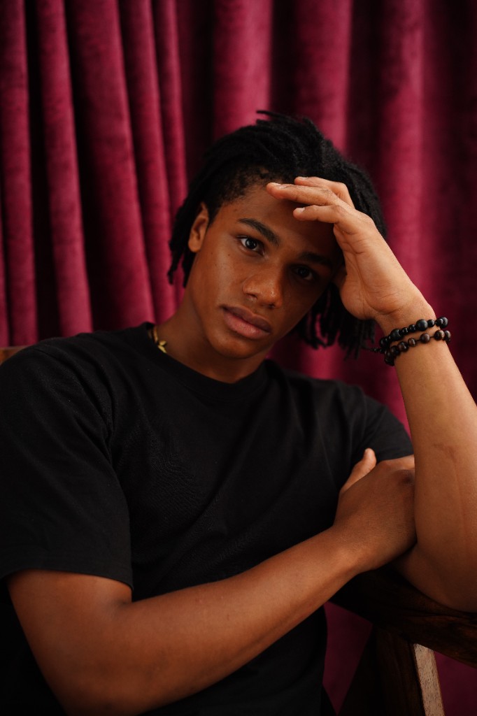 Editorial portrait of Nnamdi Unamadu seated against deep burgundy curtains